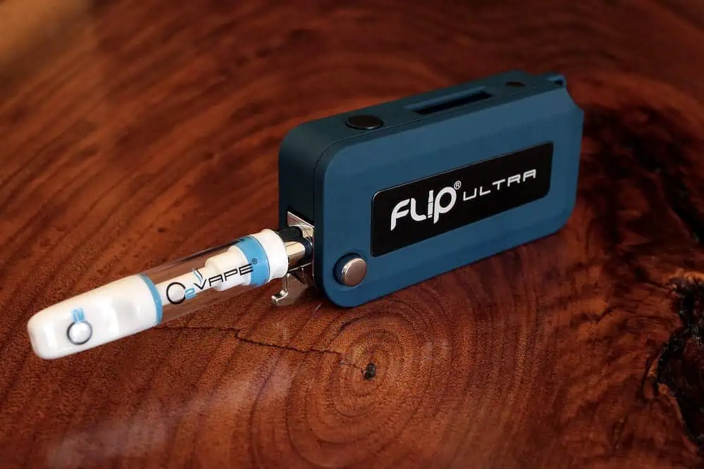 A blue and black Flip Ultra Cannabis Vape battery lies on a polished wooden surface, with a white and blue C-Vape cartridge attached. The device, featured in our Vape Hardware Guide, is rectangular with a logo and button, ready for use.