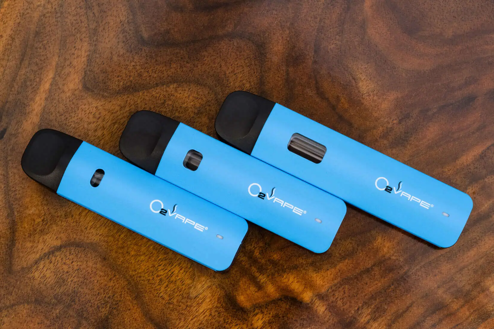 Three blue vape pens with black mouthpieces are arranged diagonally on a wooden surface. Ideal for cannabis professionals, each device displays "O2VAPE" and features a unique size and window design for checking vape liquid levels.