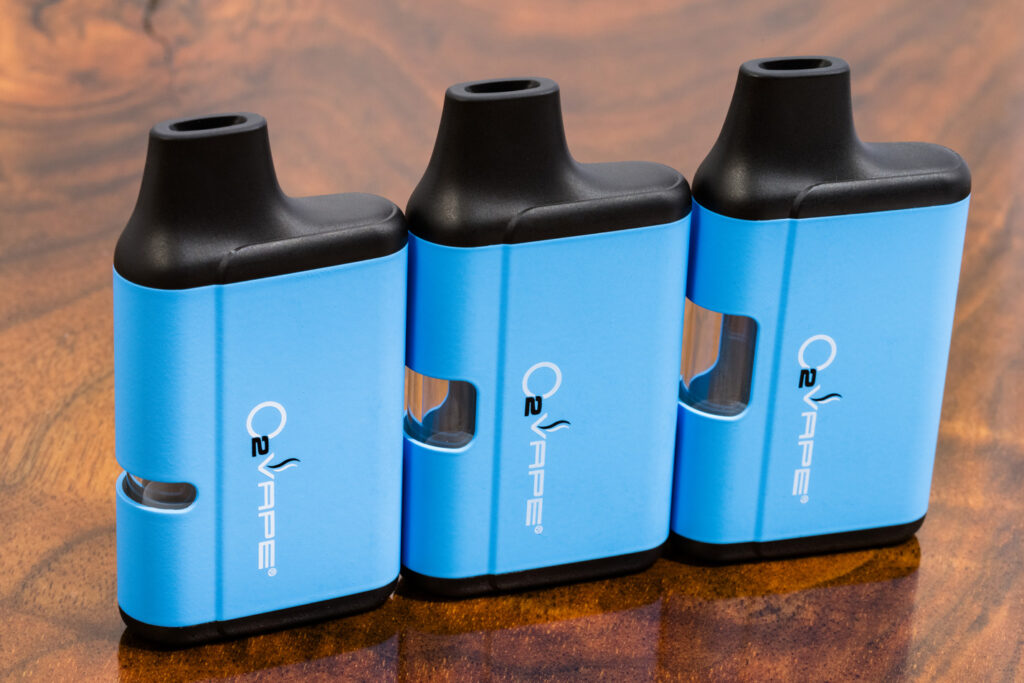 Three blue and black O2VAPE devices stand upright in a row on a wood surface. Each Cannabis Vape Hardware device features a small window revealing the cartridge, rounded edges, and the brand logo “O2VAPE” printed vertically on the front.
