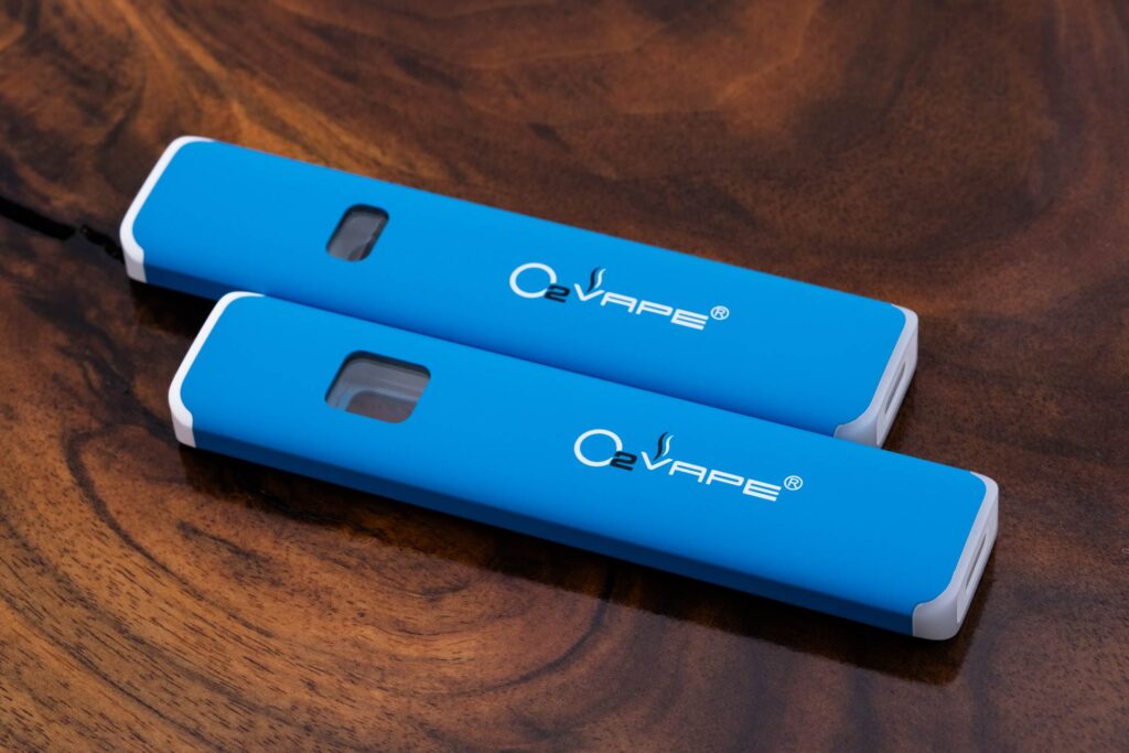 Two blue rectangular vape pens with white edges and "O2VAPE" branding lie on a wooden surface. These sleek examples of cannabis vape hardware feature small rectangular windows and silver tips, contrasting beautifully with the swirling wood grain beneath.
