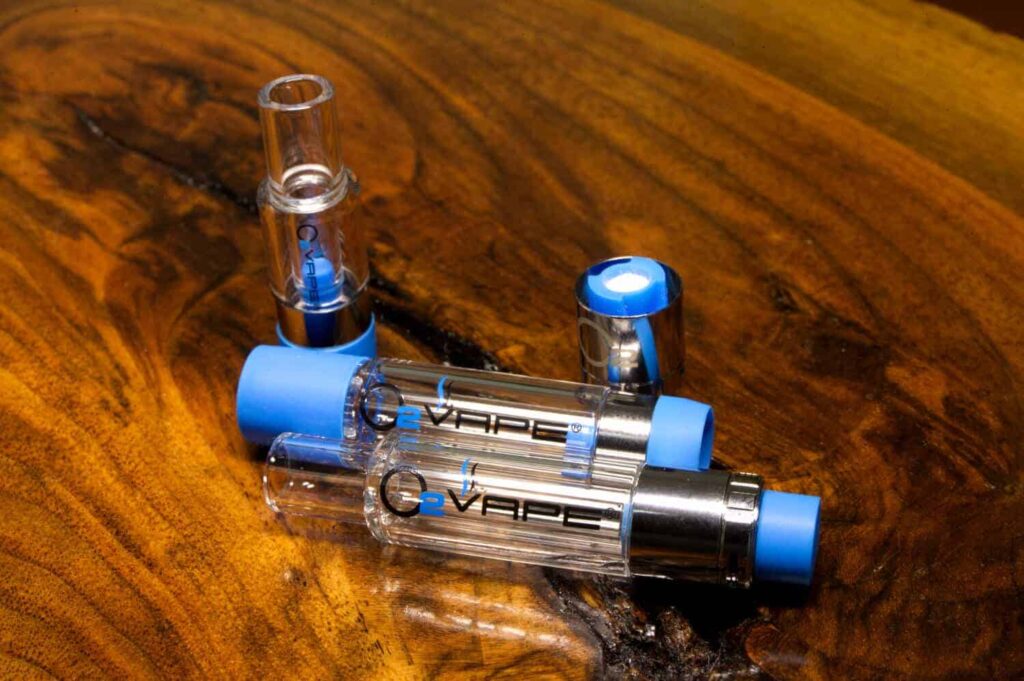 Three Q Vape live resin vape cartridges with blue tips rest on a polished, brown wooden surface. Two cartridges lie horizontally, one is upright, and a silver cap with a blue top is nearby. The logo “Q Vape” is visible on the clear glass cartridges.
