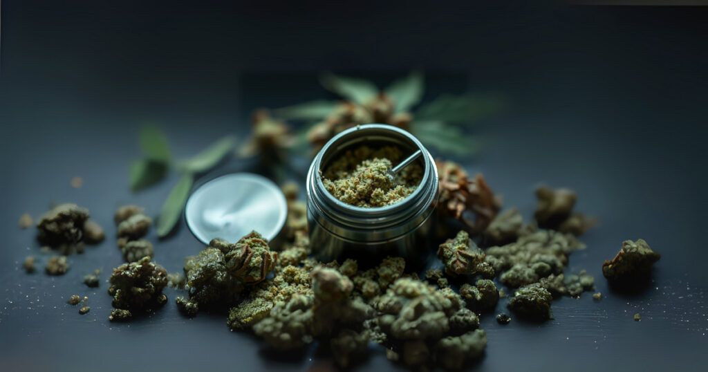 Close-up of cannabis buds scattered on a dark surface, with a small metal container at the center, its lid open and filled with ground cannabis. Some green leaves and dried plant parts are blurred in the background. The lighting is soft and dramatic.