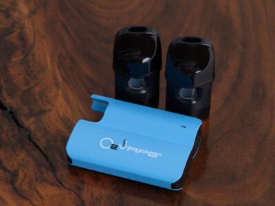 A blue nanopod O2VAPE device with the logo visible lies on a brown wood surface. Behind it are two black vape cartridges standing upright, each with clear chambers. The surface has a natural grain and a dark crevice running through it.