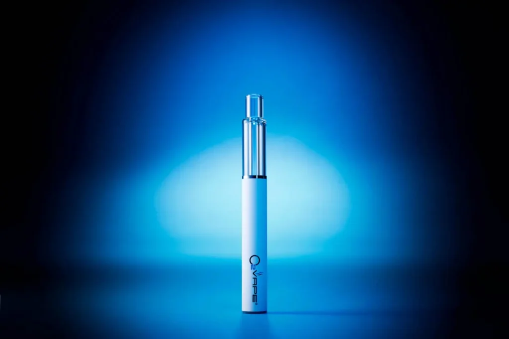 A metallic vape pen stands upright, centered against a blue gradient background. The sleek O2VAPE hardware features a silver and white, cylindrical design with a clear mouthpiece on top.